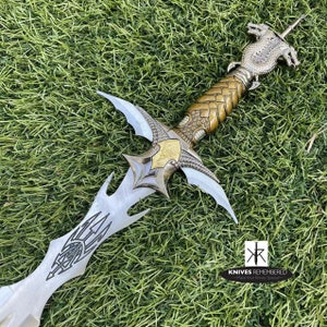 May include: A silver and gold fantasy sword with a dragon head pommel. The sword has a double-edged blade with intricate details and a black leather grip.