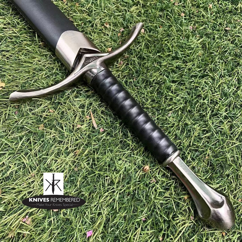 May include: A silver and black sword with a leather grip and a cross guard. The sword is lying on a bed of green grass.
