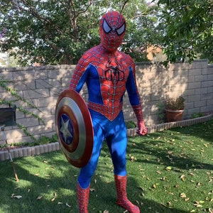 May include: A person dressed as Spider-Man is holding Captain America's shield. The shield is red, white, and blue with a star in the center. The person is standing on a green lawn.