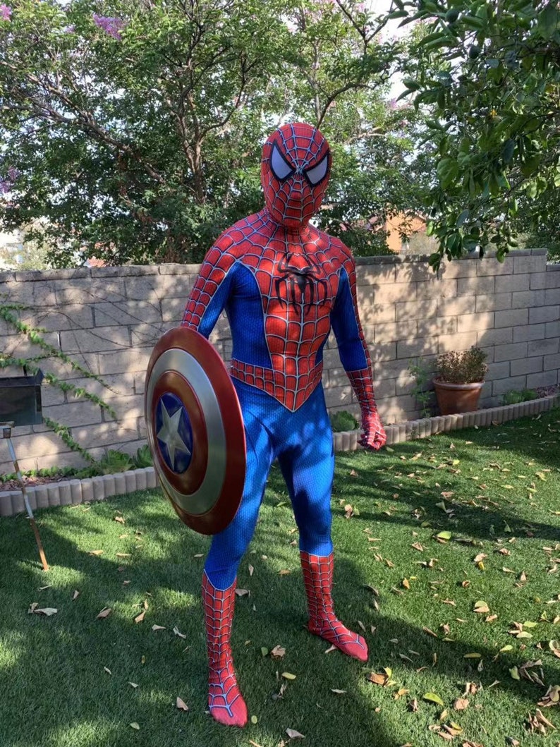 May include: A person dressed as Spider-Man is holding Captain America's shield. The shield is red, white, and blue with a star in the center. The person is standing on a green lawn.