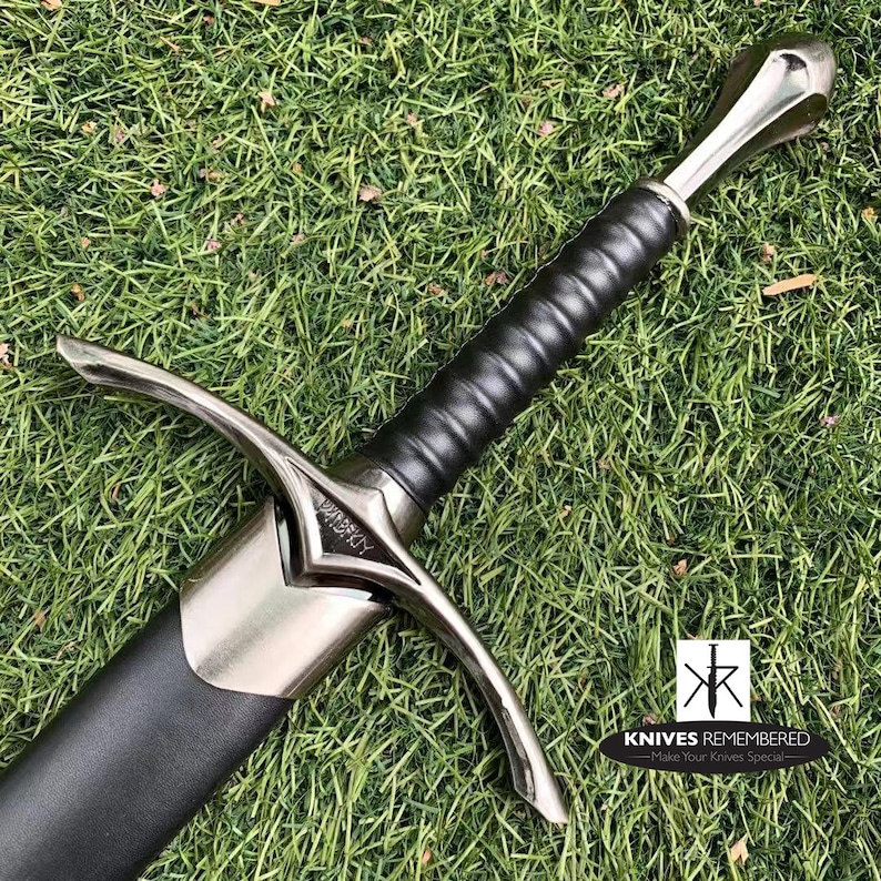May include: A silver sword with a black leather grip and a silver pommel. The sword has a crossguard with a decorative design and the word "WINDRUN" inscribed on the blade. The sword is lying on a bed of green grass.
