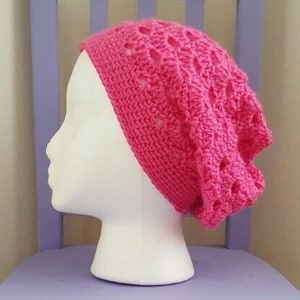 May include: A bright pink crocheted beanie hat on a white mannequin head, displayed on a lavender-colored wooden chair. The hat has an openwork design. The chair is in a room with beige walls and a carpeted floor. The text "The Lavender Chair" is at the bottom.