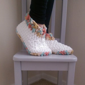 May include: White crocheted slippers with a colourful crocheted trim around the top. The slippers are sitting on a light grey chair.