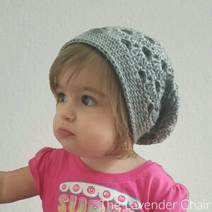 May include: A young child wearing a gray crocheted beanie with a lacy pattern. The beanie is slouchy and sits low on the forehead. The child is wearing a pink shirt with the word "SUPER" printed on it.