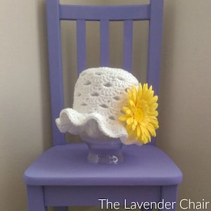 May include: White crochet sun hat with a yellow flower accent. The hat is sitting on a purple chair.