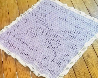 Könnte beinhalten: Eine Babydecke aus lilafarbenem Häkelgarn mit einem weißen Häkelrand. Die Decke hat ein zartes, durchbrochenes Muster.