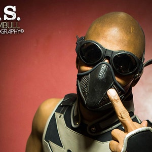 May include: A person wearing a black leather face mask with a silver nose guard and goggles. The person is wearing a black and silver vest and has a finger pointed up.
