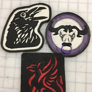 May include: Three leather patches with embroidered designs. The first patch is white with a black crow design. The second patch is purple with a white bull design. The third patch is black with a red dragon design.