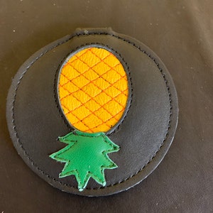 May include: A black leather keychain with a yellow pineapple design. The pineapple has a green top and is stitched with orange thread.