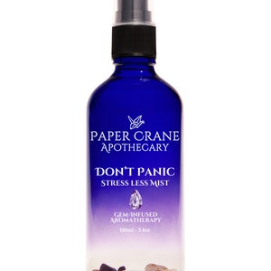 May include: A blue glass spray bottle with a black nozzle, containing small gemstones at the bottom. The label reads: PAPER CRANE APOTHECARY, DON'T PANIC STRESS LESS MIST, GEM-INFUSED AROMATHERAPY, 100ml - 3.4oz. The bottle features a gradient design.
