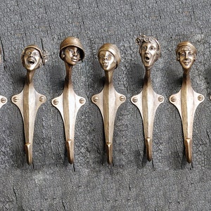 Puede incluir: Siete ganchos de pared decorativos hechos de metal con un acabado dorado. Cada gancho presenta una cara &uacute;nica esculpida en el metal. Los ganchos est&aacute;n montados en una superficie de madera.