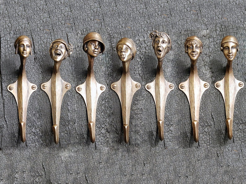 Puede incluir: Siete ganchos de pared decorativos hechos de metal con un acabado dorado. Cada gancho presenta una cara &uacute;nica esculpida en el metal. Los ganchos est&aacute;n montados en una superficie de madera.