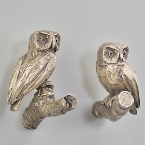 May include: Two metallic, bronze-toned wall hooks, each shaped like an owl perched on a textured tree branch. The owls have detailed, carved-looking feathers and round eyes, with a weathered, antique finish on the metal surface.