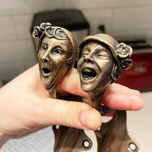 Puede incluir: Dos tiradores de puerta decorativos de bronce con forma de cabezas de mujer. Una mujer est&aacute; riendo con la boca abierta y los ojos cerrados. La otra mujer est&aacute; sorprendida con la boca abierta y los ojos muy abiertos.