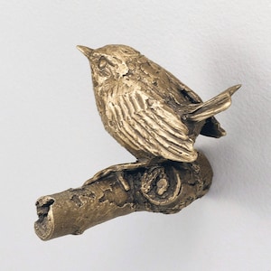May include: A metallic bronze-toned wall hook shaped like a small bird perched on a textured tree branch. The bird features detailed wing feathers and a pointed beak, while the branch has a rough, bark-like surface texture throughout the design.