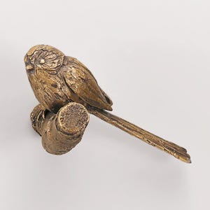 May include: A bronze-colored metal figurine of a small bird with textured plumage perched on a cylindrical branch. The bird has a rounded head, a short beak, and a long, slender tail extending horizontally from the branch, showing a metallic finish.