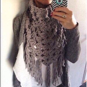 May include: A grey crocheted scarf with fringe. The scarf is worn around the neck and has a triangular shape.