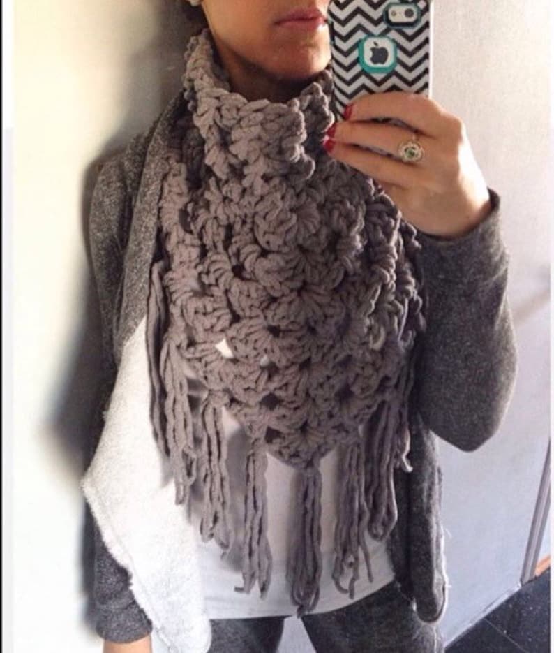 May include: A grey crocheted scarf with fringe. The scarf is worn around the neck and has a triangular shape.