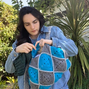 May include: A person wearing a denim jacket is holding a crocheted pet carrier with blue and grey squares. The carrier is made of yarn and has a soft, cosy texture.
