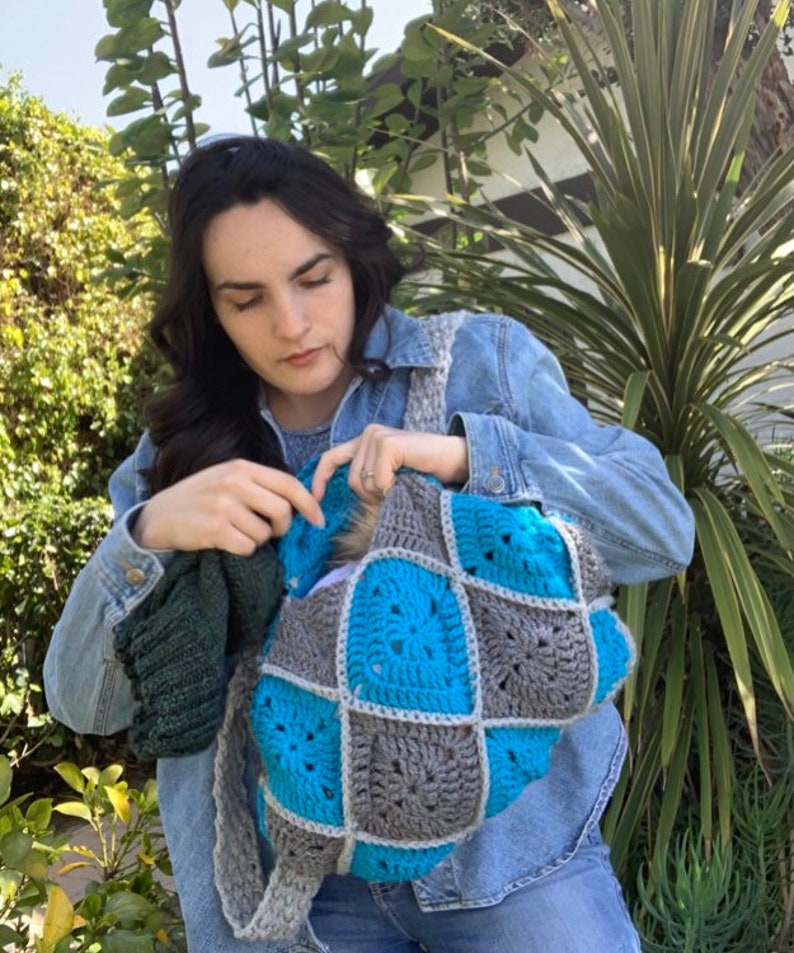 May include: A person wearing a denim jacket is holding a crocheted pet carrier with blue and grey squares. The carrier is made of yarn and has a soft, cosy texture.