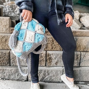May include: A crocheted tote bag with a blue and grey checkered pattern. The bag has a tassel and a long strap. The bag is being held by a person wearing black jeans and a plaid shirt.