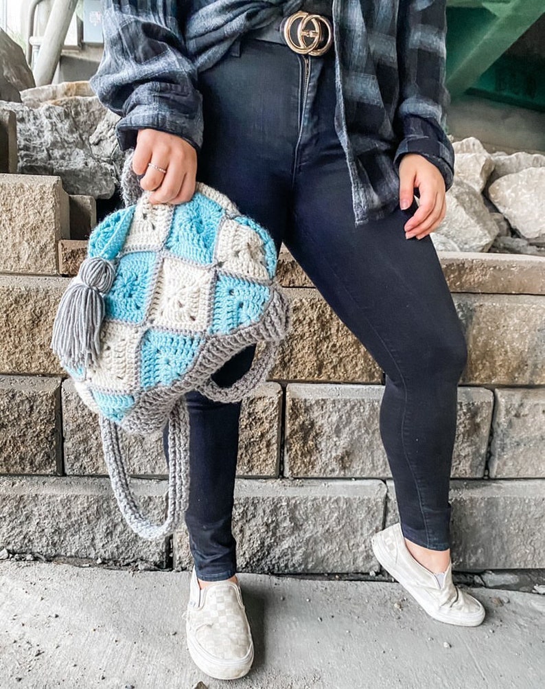 May include: A crocheted tote bag with a blue and grey checkered pattern. The bag has a tassel and a long strap. The bag is being held by a person wearing black jeans and a plaid shirt.