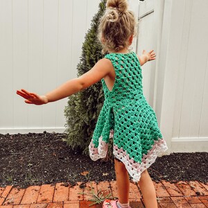 May include: A green crochet dress with a white and pink border. The dress is sleeveless and has a loose, open weave design. The child is wearing pink clogs with a cartoon design.