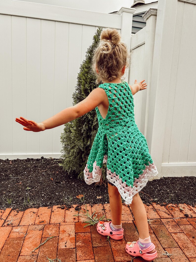 May include: A green crochet dress with a white and pink border. The dress is sleeveless and has a loose, open weave design. The child is wearing pink clogs with a cartoon design.