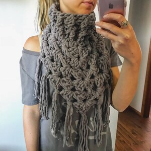 May include: A grey crocheted scarf with fringe. The scarf is worn around the neck of a person.