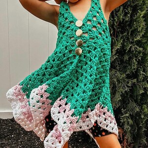May include: A green crochet dress with a V-neck and gold buttons down the front. The dress has a scalloped hemline with white and pink accents. The dress is handmade and suitable for a child.