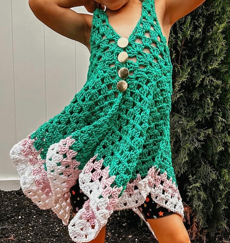 May include: A green crochet dress with a V-neck and gold buttons down the front. The dress has a scalloped hemline with white and pink accents. The dress is handmade and suitable for a child.
