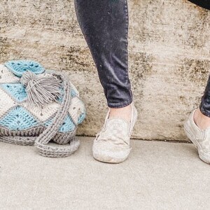 May include: A crocheted bag with a blue and white diamond pattern and a grey tassel. The bag is lying on a concrete surface.