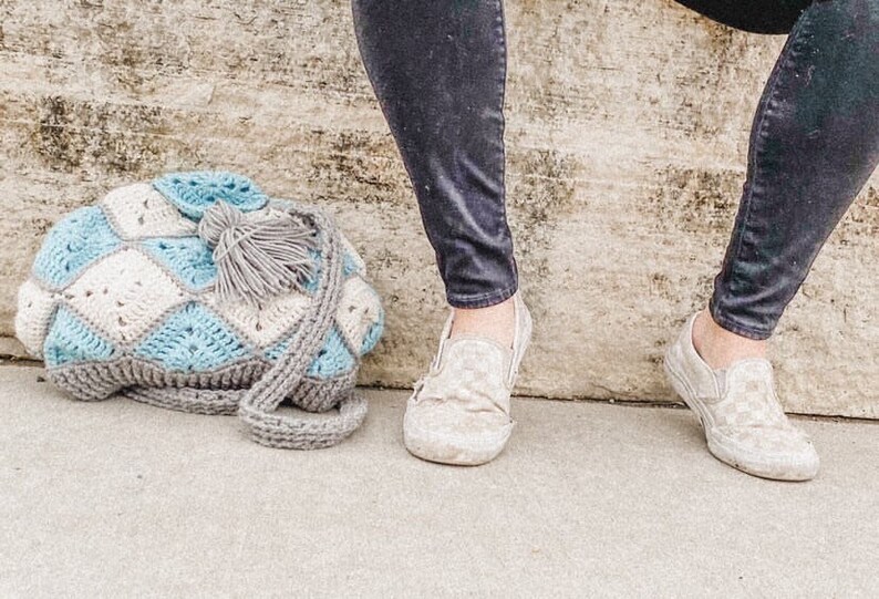May include: A crocheted bag with a blue and white diamond pattern and a grey tassel. The bag is lying on a concrete surface.