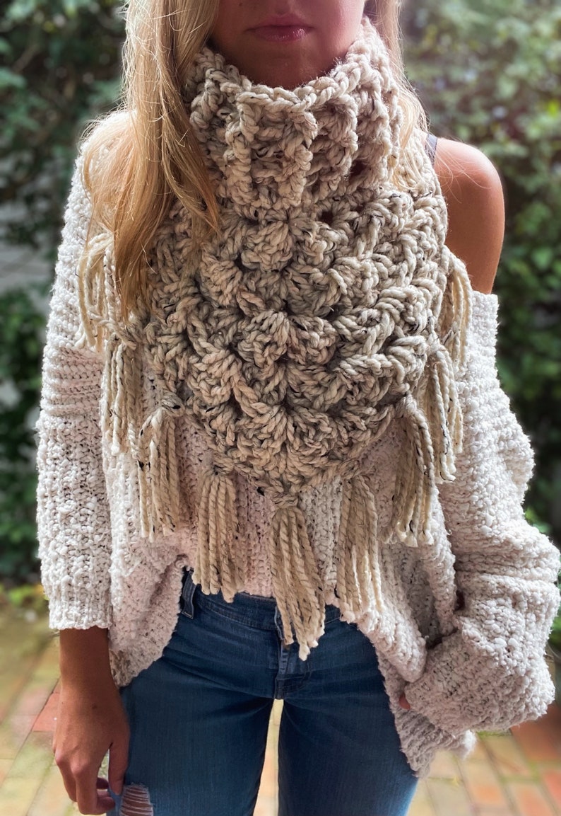 May include: A crocheted cream-coloured scarf with fringe. The scarf is made with a textured stitch and has a triangular shape.