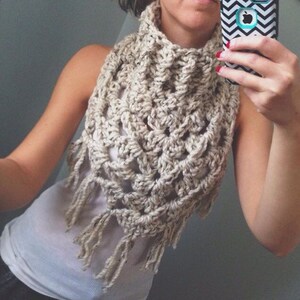 May include: A crocheted cowl with fringe in a cream colour. The cowl is worn by a person and is shown from the front.
