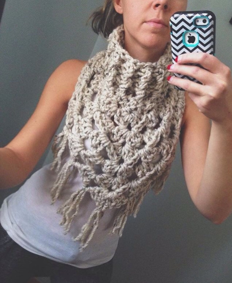 May include: A crocheted cowl with fringe in a cream colour. The cowl is worn by a person and is shown from the front.