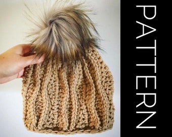 Op de afbeelding: Een gebreide beanie muts in een lichtbruine kleur met een pluche bruine en zwarte pompon bovenop. De muts heeft een geribbelde textuur.