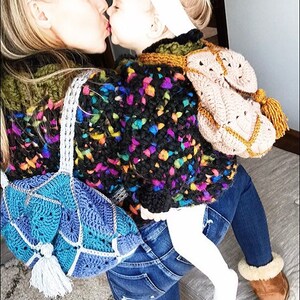 May include: A woman and a young child wearing colourful crocheted sweaters. The woman is wearing blue jeans and a blue crocheted bag. The child is wearing a white headband and white trousers. They are kissing each other.