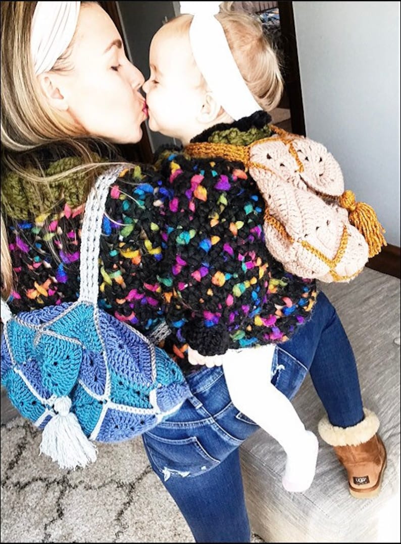 May include: A woman and a young child wearing colourful crocheted sweaters. The woman is wearing blue jeans and a blue crocheted bag. The child is wearing a white headband and white trousers. They are kissing each other.
