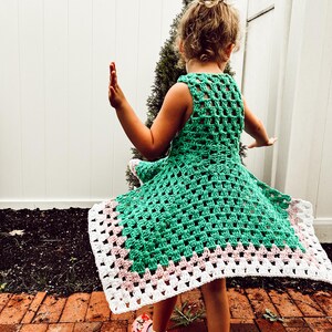 May include: A green crochet dress with a white and pink border. The dress is sleeveless and has a square pattern. The dress is being worn by a person with their arms outstretched. The person is wearing pink clogs.