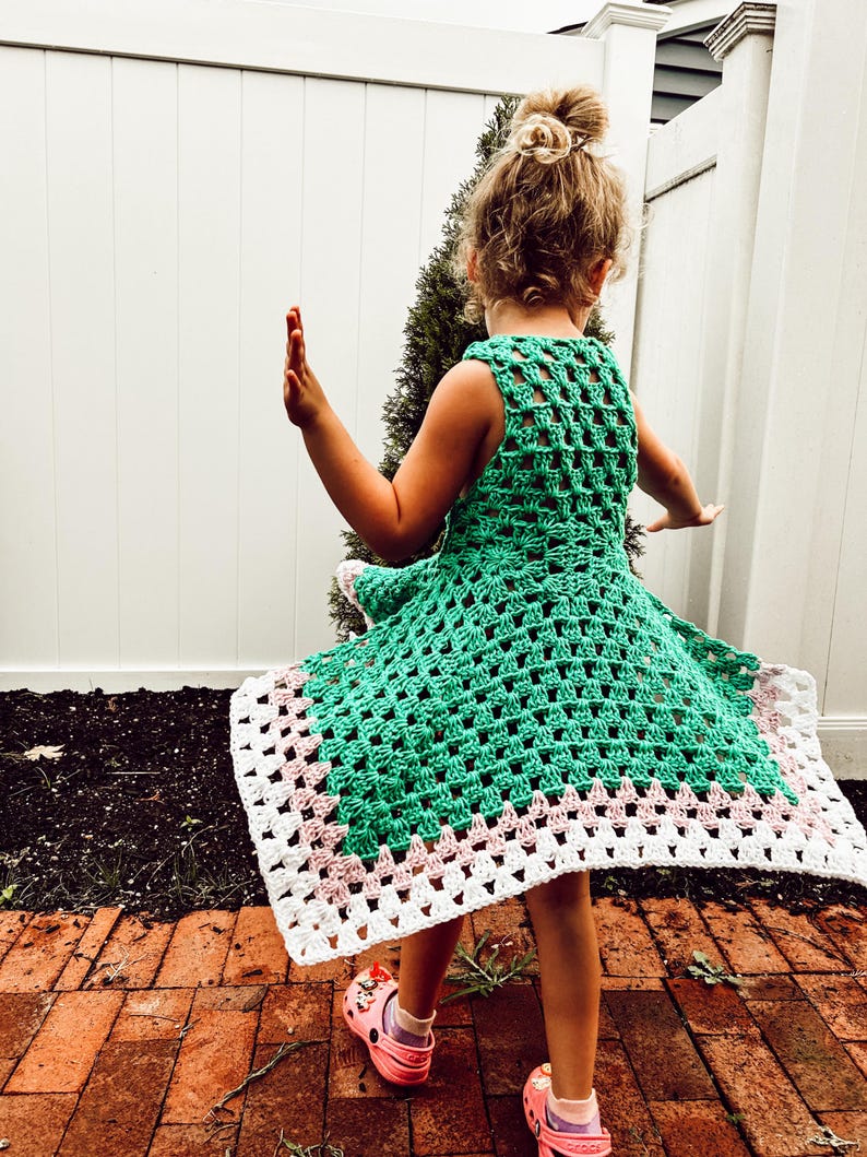 May include: A green crochet dress with a white and pink border. The dress is sleeveless and has a square pattern. The dress is being worn by a person with their arms outstretched. The person is wearing pink clogs.