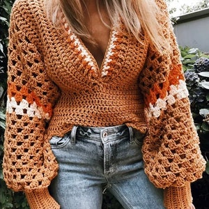 May include: A brown crochet top with a V-neck and long sleeves. The top has a white and orange stripe pattern around the neckline and cuffs. The top is worn with blue jeans.