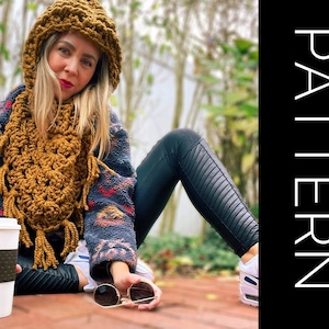 May include: A woman wearing a brown crocheted hat and scarf with fringe. She is sitting on a brick surface with a cup of coffee and sunglasses.