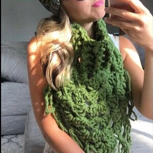 May include: A woman wearing a green crocheted scarf with fringe and a green, blue, and brown crocheted beanie with a faux fur pom pom. She is also wearing black cat eye sunglasses.