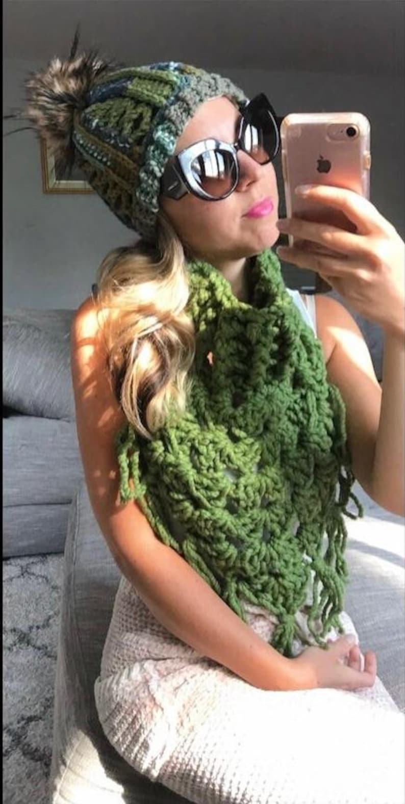 May include: A woman wearing a green crocheted scarf with fringe and a green, blue, and brown crocheted beanie with a faux fur pom pom. She is also wearing black cat eye sunglasses.