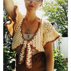 May include: A crocheted crop top with a tie front and ruffled sleeves. The top is made in a cream and brown color scheme.