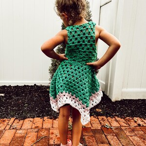 May include: A green crochet dress with a white and pink trim. The dress is sleeveless and has a flared skirt. The child is wearing pink sandals. The background includes a white fence and a brick patio.