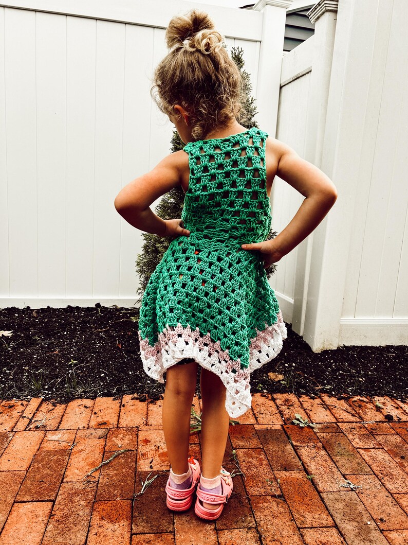May include: A green crochet dress with a white and pink trim. The dress is sleeveless and has a flared skirt. The child is wearing pink sandals. The background includes a white fence and a brick patio.