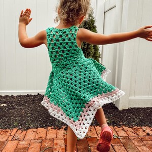May include: A green crochet dress with a white and pink border. The sleeveless dress is made of a open weave pattern. The dress is worn by a person wearing pink shoes.