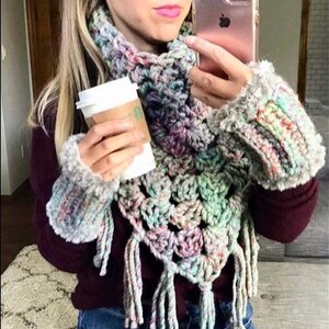 May include: A woman wearing a multi-coloured crochet scarf and matching arm warmers with long tassels. She is also wearing a maroon sweater and blue jeans.